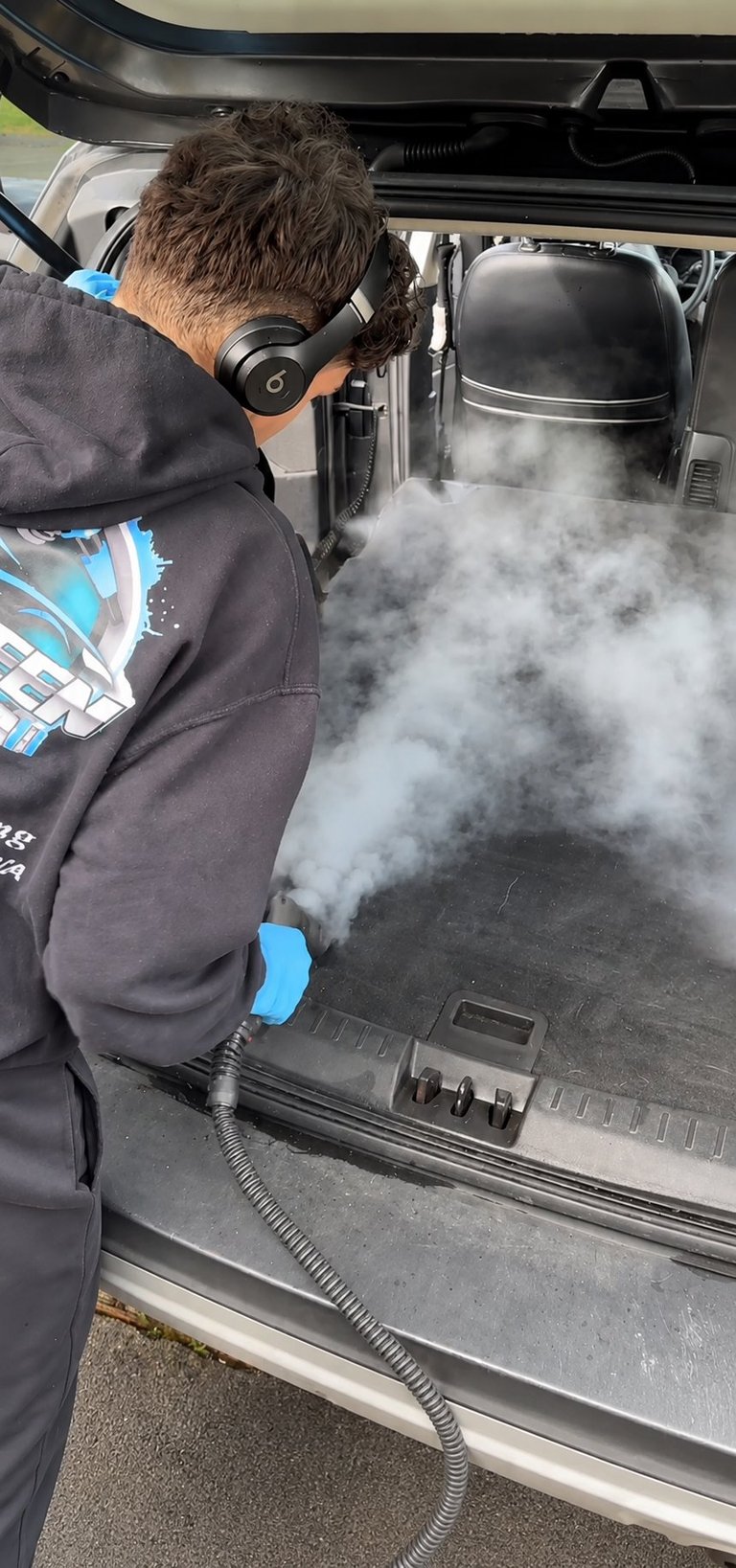 Person using dry ice cleaning equipment on car trunk interior, producing heavy white vapor