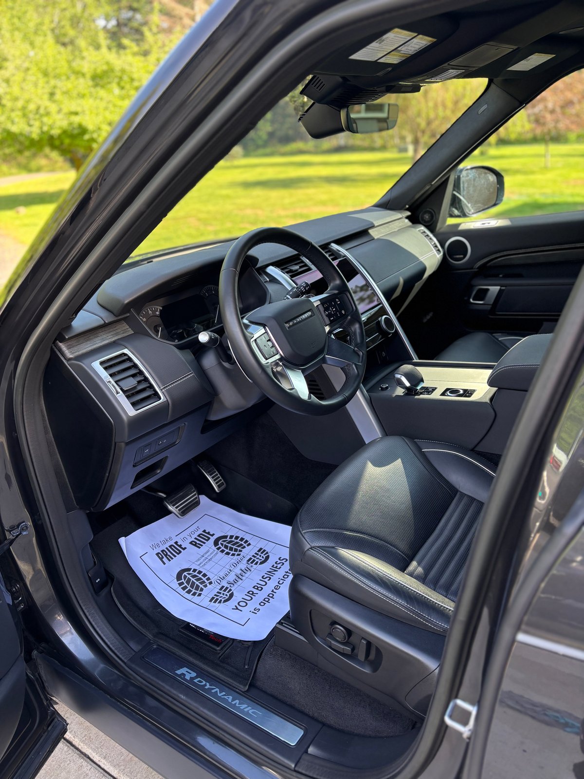 Interior view of a luxury SUV with steering wheel, dashboard, and green landscape visible through the open driver door